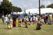 Thumbnail for Children participating in a sack race on a grassy field during a sunny outdoor community event with tents and spectators.