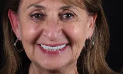 Thumbnail control image for Smiling mature woman with brown hair, wearing hoop earrings and a black top against a black background.