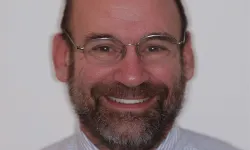 Thumbnail control image for Close-up portrait of a smiling middle-aged man with glasses, beard, and striped shirt against a plain background