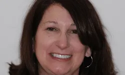 Thumbnail control image for Smiling middle-aged woman with shoulder-length brown hair wearing silver hoop earrings against a plain background