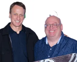 Two men posing with a Tony Hawk Birdhouse skateboard featuring a bird skull graphic on wood grain.