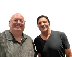 Two smiling men posing together against a white background, one with glasses and striped shirt, the other in black t-shirt.