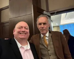 Two men posing for a photo indoors, one smiling in a black blazer and the other serious in a brown suit and tie.