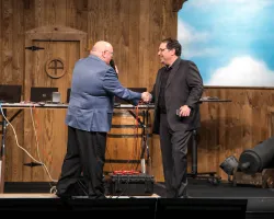 Two men shaking hands on a stage with wooden backdrop and vintage cannon in the background