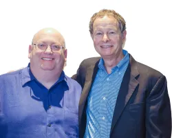Two middle-aged men smiling, one wearing glasses and a blue shirt, the other in a blue checkered shirt and black blazer.