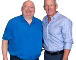 Two men standing side by side smiling, one in a blue shirt and the other in a light blue button-down shirt against white background