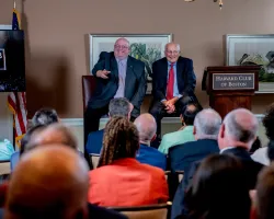 Two men speaking to an audience at Harvard Club of Boston during a formal event or summit.