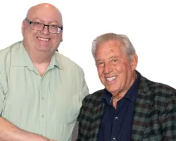 Two men smiling and shaking hands, one standing in light green shirt and jeans, the other seated in plaid jacket and brown pants