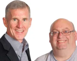 Two men smiling at the camera against a white background, one in a suit and the other in a casual shirt and glasses