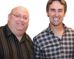 Two men smiling, one bald wearing glasses and a striped shirt, the other with brown hair in a plaid shirt