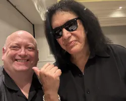 Two men smiling indoors, one wearing sunglasses and black shirt, the other pointing to him with a thumb up gesture.