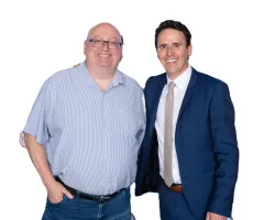 Two men smiling, one in a blue striped shirt and jeans, the other in a navy suit and beige tie, standing against white background