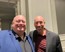 Two smiling men in business casual attire standing indoors against a light-colored wall with molding.