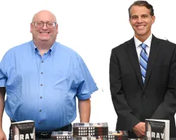 Two men smiling behind a table with stacks of the book titled Gray Day on a white background.