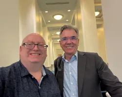 Two smiling men wearing glasses posing for a selfie in a well-lit hallway with white walls and ceiling lights.