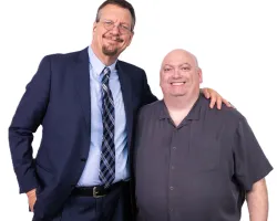 Two men smiling with one in a suit and tie and the other in a casual shirt against a white background