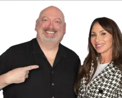 Smiling man in black shirt points to smiling woman in black and white houndstooth blazer against white background