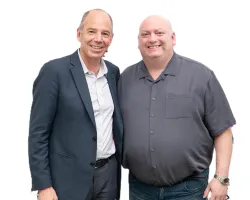 Two middle-aged men smiling, one in a suit and the other in a casual shirt, against a white background.