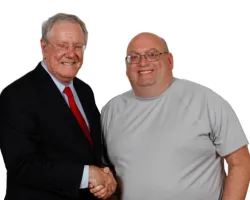 Two men smiling and shaking hands; one in a suit with red tie, the other in a gray t-shirt and glasses