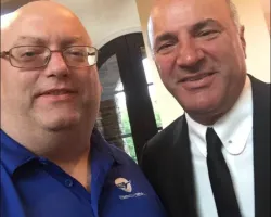 Two middle-aged men posing for a close-up photo indoors, one in glasses and blue shirt, the other in a black suit and tie.