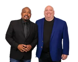 Two men smiling, one in a black suit and tie, the other in a blue blazer and black shirt, standing on white background