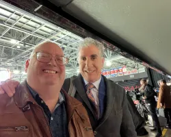 Two men smiling in a large indoor arena with bright lights and a crowd in the background.