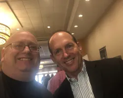 Two men smiling indoors at a social event with warm lighting and people in the background.