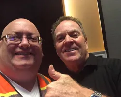 Two smiling men, one in safety vest and glasses, the other giving thumbs up, indoors with warm lighting.