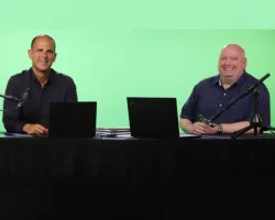 Two men sitting at a desk with laptops and microphones in front of a green screen background.
