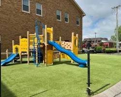 Yellow and blue playground structure with slides and ladders on green artificial grass near brick building.