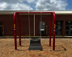 Empty black basket swing with red frame on wood chips next to brick school building on a partly cloudy day