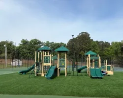 Playground with green slides and climbing structures on artificial grass surrounded by trees and blue sky.