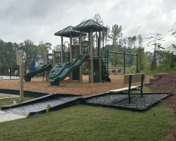 Outdoor playground with slides, swings, ramp access, and a bench surrounded by trees and neighborhood homes.
