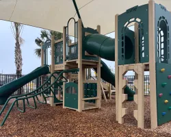 Modern playground with slides, climbing structures, and monkey bars under a shaded canopy on wood chip ground.