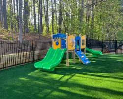 Children's playground with green slides, climbing structure, and artificial grass surrounded by trees and a fence