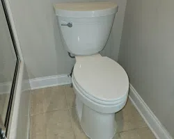 White porcelain toilet with closed lid in a beige tiled bathroom corner next to glass shower door.
