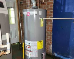 Gray Rheem water heater with energy guide label installed in a basement with brick wall and blue painted section.