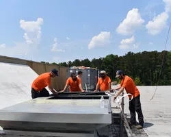 Five workers in orange shirts install or maintain rooftop HVAC equipment under a sunny blue sky.