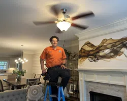 Handyman in orange shirt standing on ladder in living room with modern decor and ceiling fan spinning above.