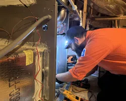 Technician in an orange shirt repairing HVAC system in a basement with headlamp light.