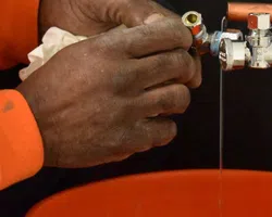 Close-up of a person’s hands fixing a valve on copper plumbing pipes over an orange bucket.