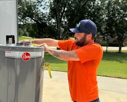 Technician in orange shirt installing or inspecting a Rheem water heater outdoors on a sunny day.