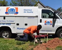 Technician in orange shirt working on plumbing near Estes Services van in suburban neighborhood.