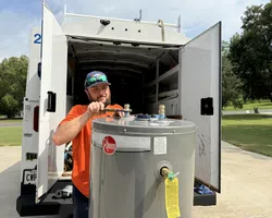 Technician in orange shirt working on a Rheem water heater outside an open service van under clear skies.