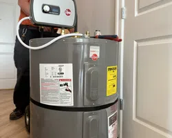 Technician in orange installing a gray Rheem water heater inside a home with wooden floor and white doors.