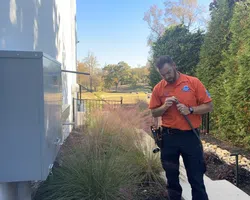 Technician in orange shirt working on outdoor electrical wiring beside a house with greenery and clear sky.