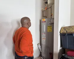 Technician in orange sweatshirt inspecting a residential ProMax water heater with pipes in a utility room.