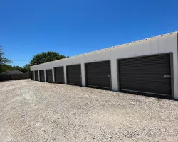 Row of white storage units with closed black roll-up doors under clear blue sky on gravel ground.