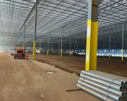 Large industrial warehouse under construction with yellow columns, steel pipes, and dirt floor visible.