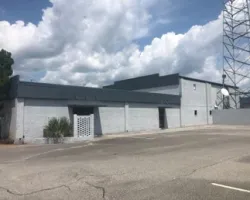Wide view of a gray industrial building with a large parking lot and a tall communication tower under cloudy sky.